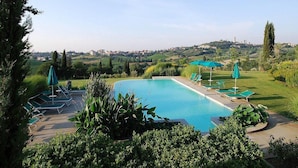 A swimming pool with lounge chairs and umbrellas, surrounded by greenery and a view of a distant town.