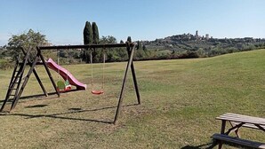A playground with a slide and swings in a grassy field with a hill and buildings in the background.