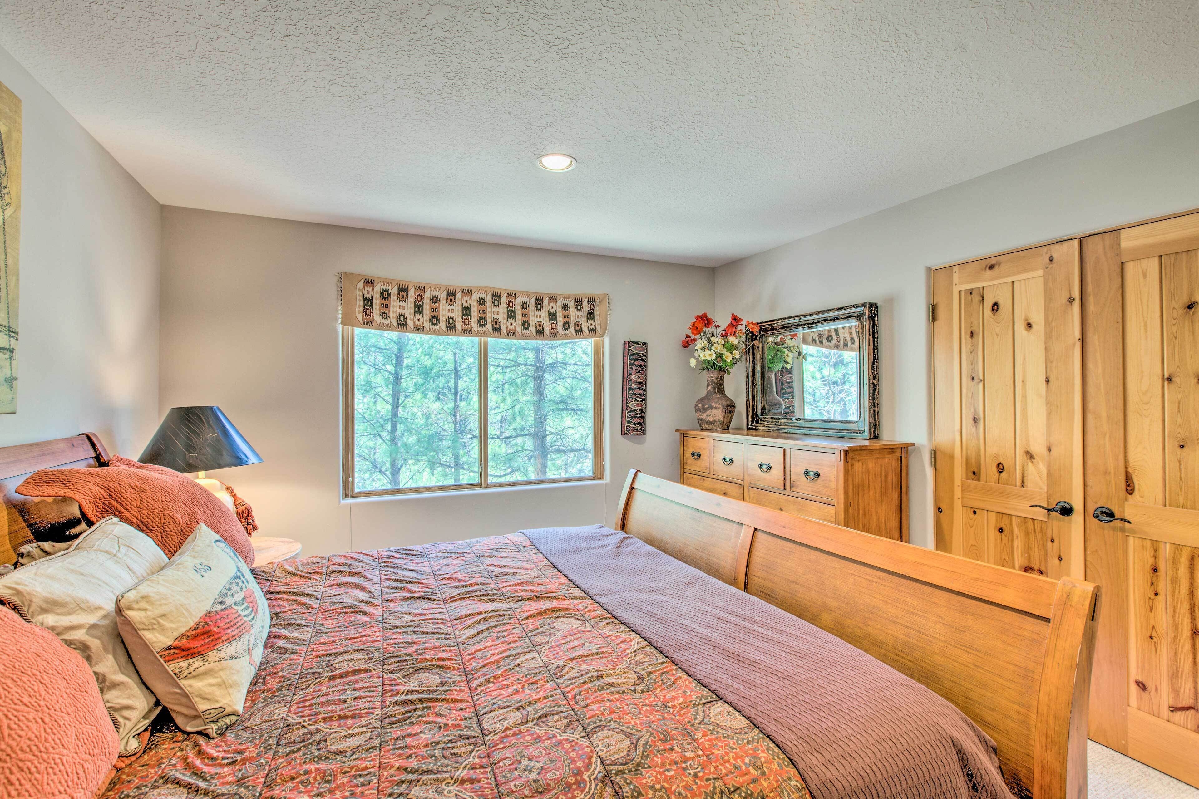 Une chambre à coucher avec un grand lit, une commode, une armoire, une fenêtre donnant sur des arbres et un luminaire au plafond.