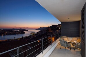 En balkon med udsigt over en kystby i skumringen, med et bord og stole.