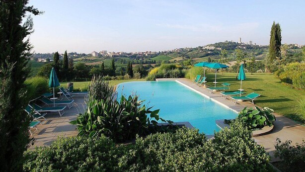 Piscina con sillones de descanso y sombrillas, rodeada de vegetación y vistas a un pueblo lejano.