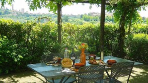 Un área de comedor al aire libre con una mesa dispuesta para dos personas, rodeada de vegetación exuberante y un paisaje urbano al fondo.