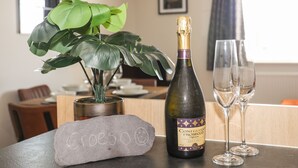 A bottle of wine, two glasses, a stone, and a plant on a table in a room with a dining area and a window. - 4 Pen Llanw Tides Reach (Rhosneigr)