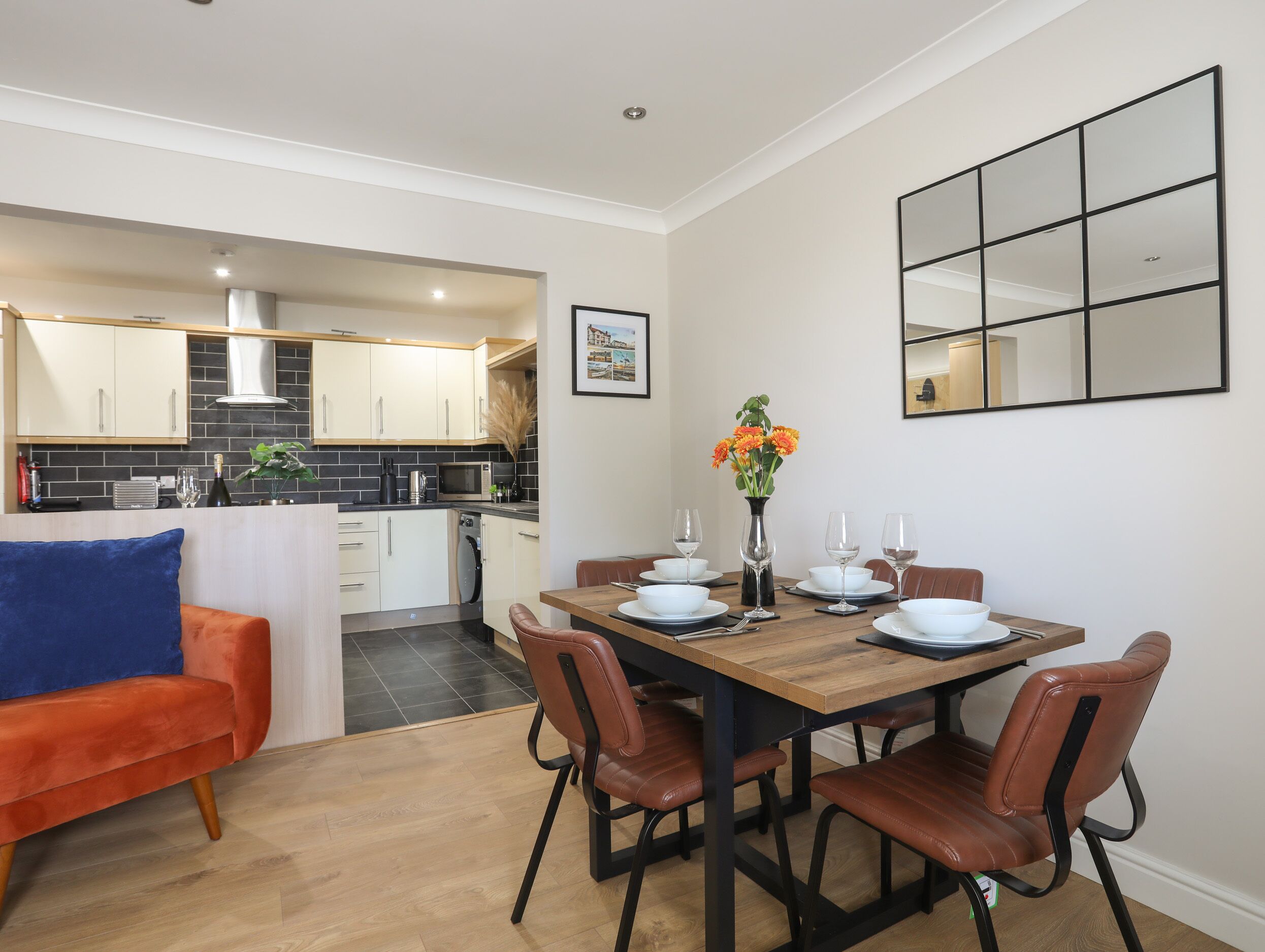 Una cocina moderna con armarios blancos, salpicadero de azulejos negros y una zona de comedor con mesa de madera y sillas de cuero marrón.