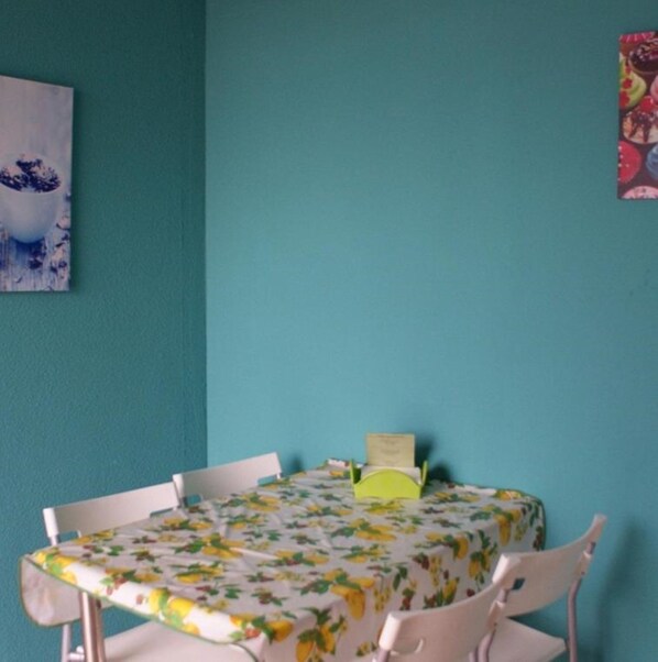 Una habitación con paredes verde azulado, una mesa con mantel floral, sillas blancas y un objeto pequeño verde sobre la mesa.