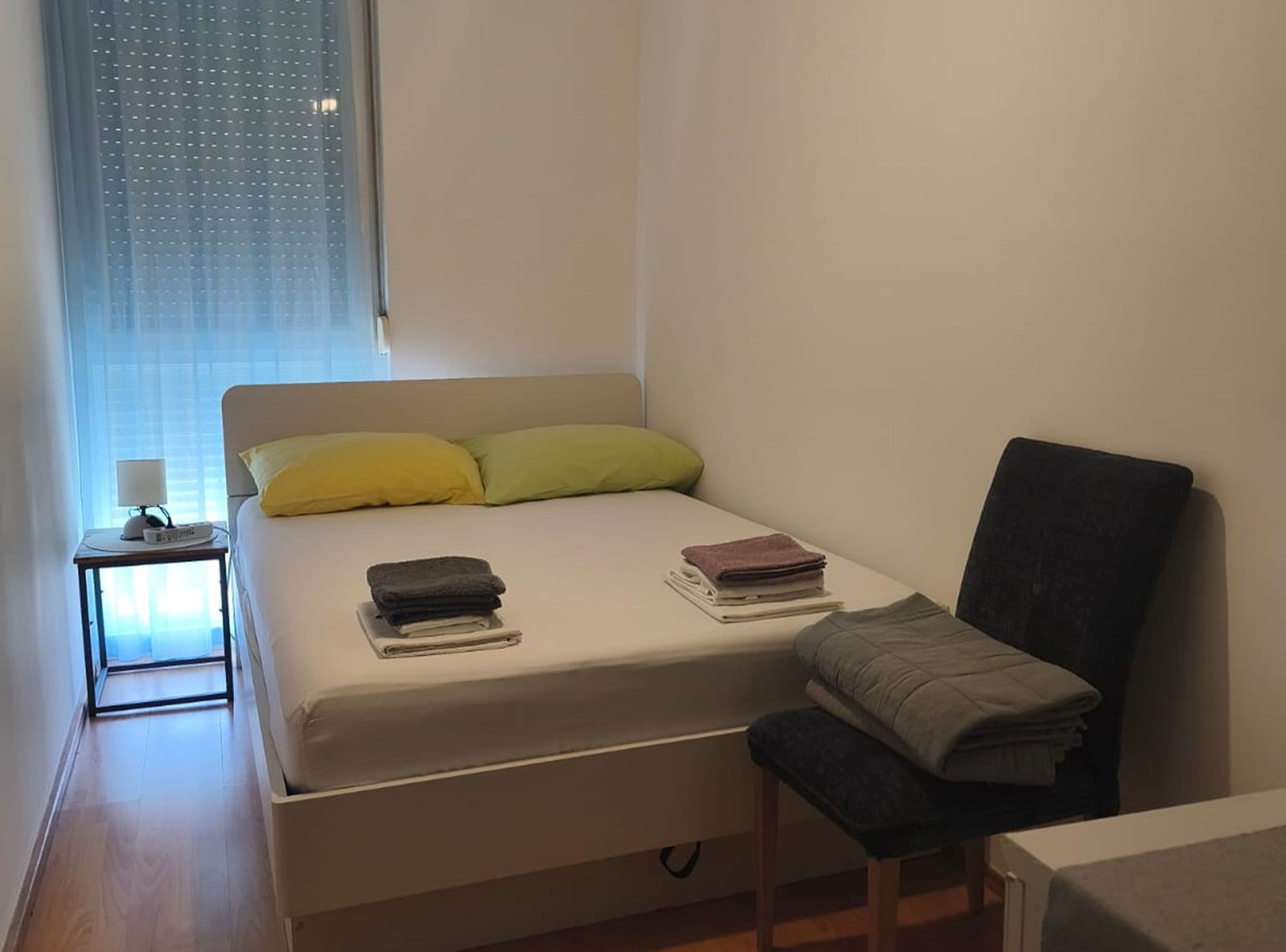 Een compacte, keurig ingerichte slaapkamer met een bed, kussens, een nachtkastje met een lamp en een stoel.