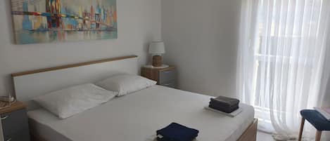A neatly made bed with white linens, a lamp on a bedside table, and a painting of a bridge on the wall.