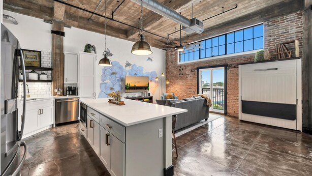 Una cocina amplia con una isla grande, armarios blancos y electrodomésticos de acero inoxidable. Hay una zona de comedor con un sofá gris y un amplio ventanal que proporciona luz natural.
