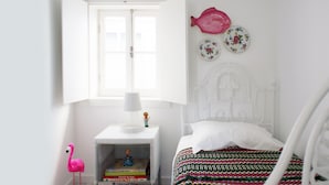 Un dormitori amb un llit blanc, una tauleta de nit amb una làmpada, una petita estant amb llibres, una decoració de flamenc rosa i una finestra amb una persiana oberta.