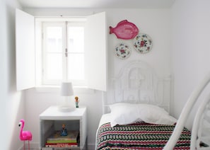 A bedroom with a white bed, a nightstand with a lamp, a small shelf with books, a pink flamingo decoration, and a window with an open shutter. - T2 - Centro Histórico da Unesco (Coimbra)