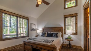 Une chambre avec un grand lit, une tête de lit en bois et deux fenêtres équipées de stores.