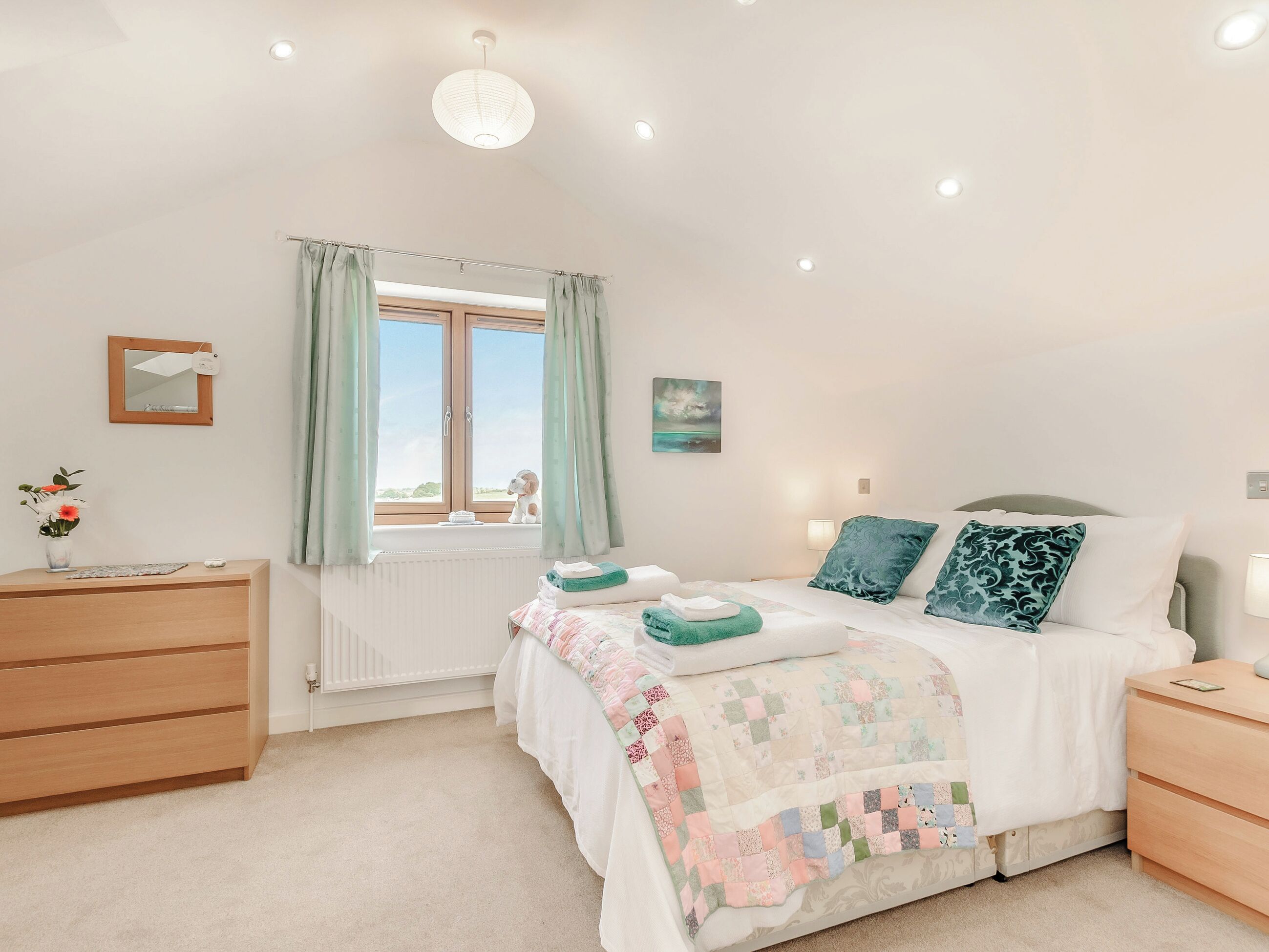 A bedroom with a bed, bedside tables, a dresser, a window with curtains, a radiator, a wall-mounted mirror, and a small painting.