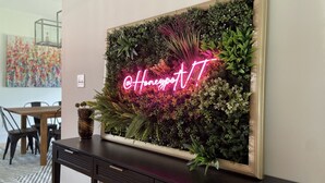 Un letrero de neón montado en la pared con '@HoneypotIT' en una habitación moderna con una consola oscura, una planta en maceta y un cuadro abstracto colorido al fondo.