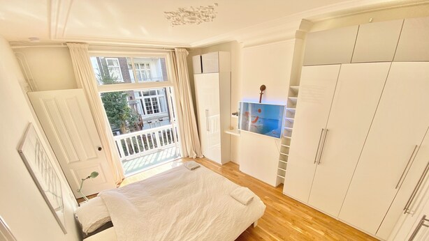 Un dormitorio con suelo de madera, cama con ropa blanca, armario y ventana con cortinas.