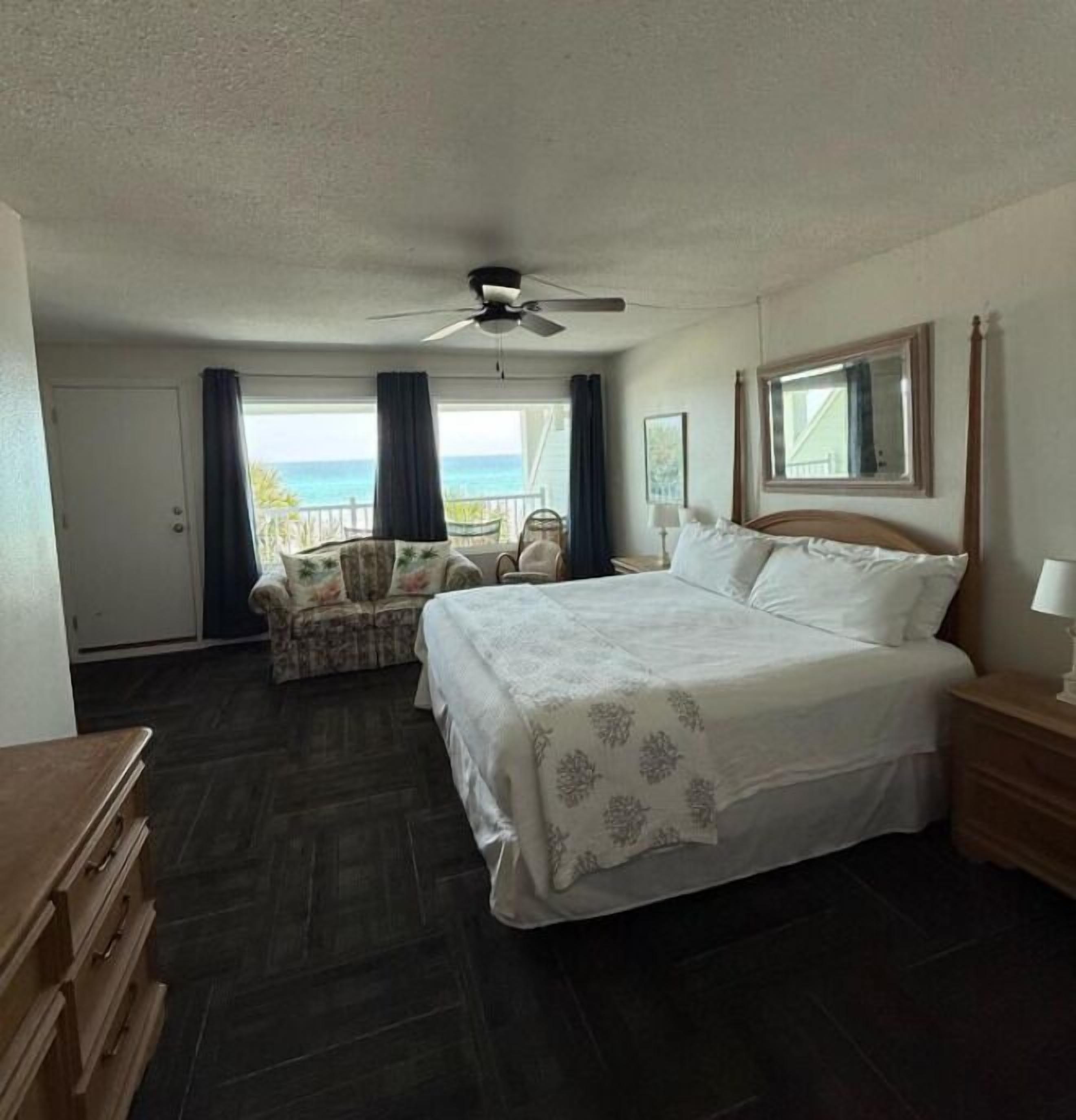A bedroom with a bed, a ceiling fan, a window with a view of the ocean, a chair, and a dresser.