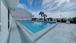A rectangular swimming pool with clear blue water, surrounded by white lounge chairs and palm trees.