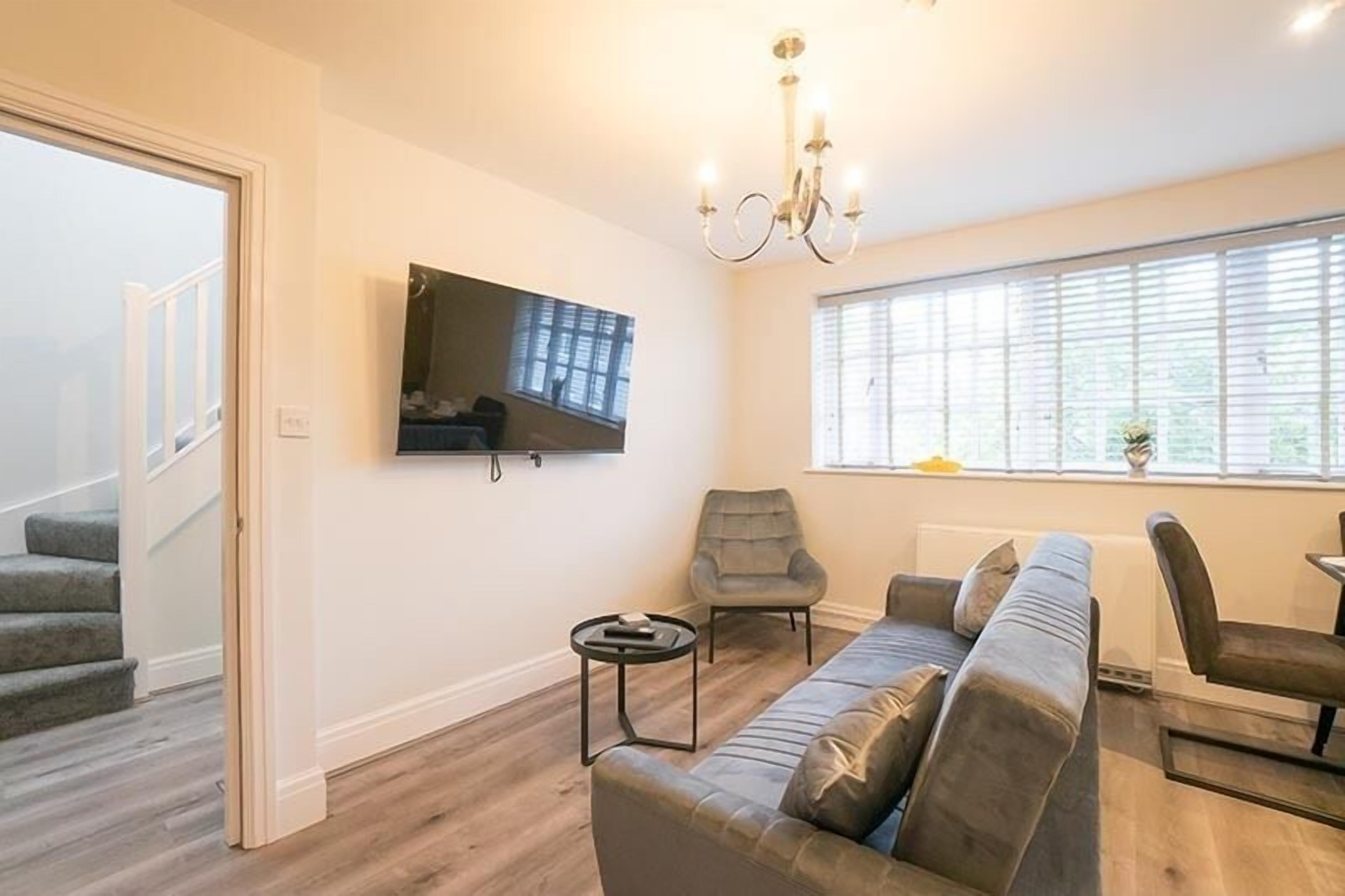 A modern living room with a grey sofa, a small round table, a grey armchair, a TV mounted on the wall, and a large window with blinds.