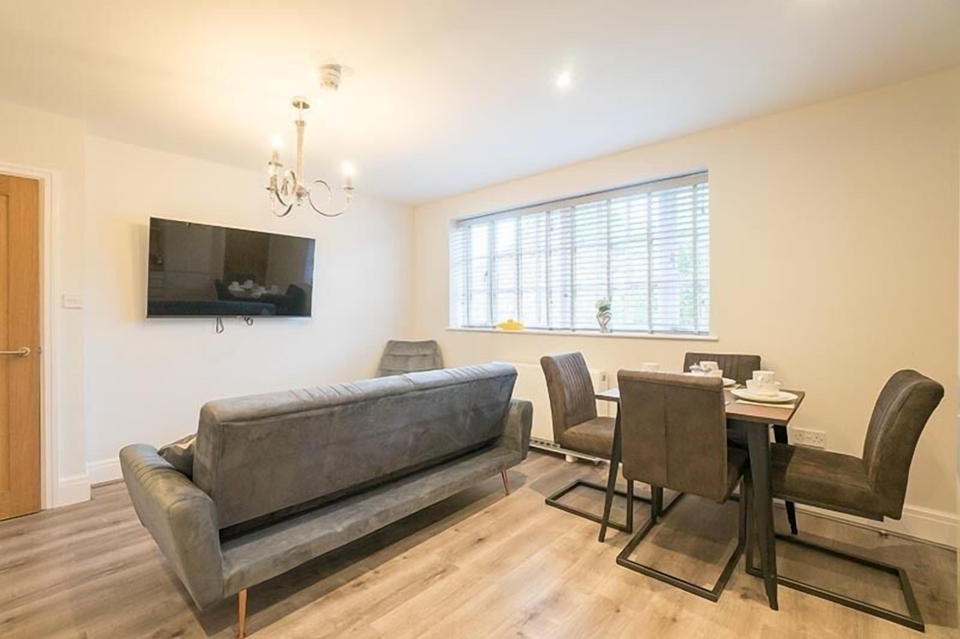 A modern living room with a grey sofa, a dining table with chairs, a flat-screen TV mounted on the wall, and a window with blinds.