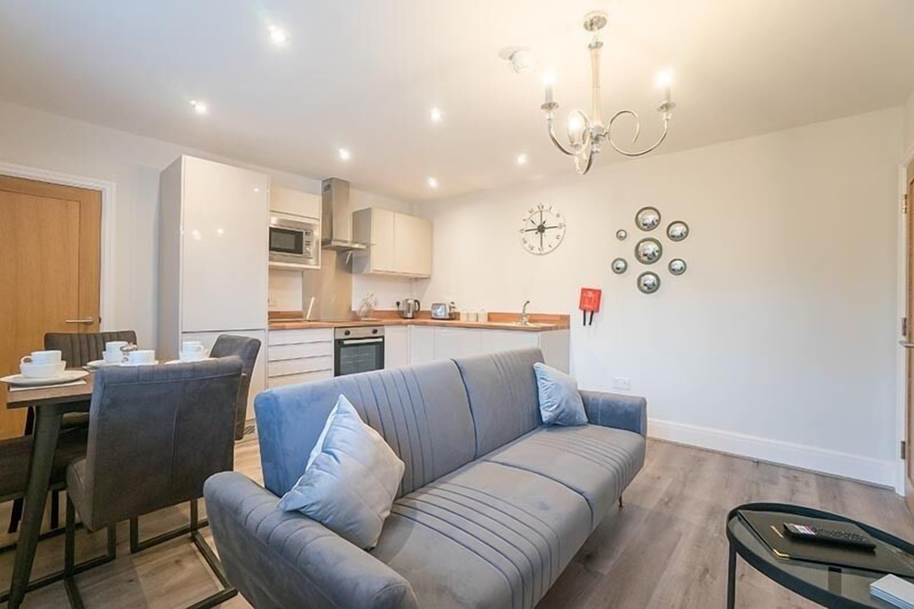 A modern living room with a grey sofa, a dining area with a table and chairs, a kitchen with white cabinets, and a chandelier.