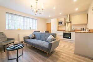 A modern apartment with a grey sofa, a round coffee table, a dining area, and a kitchen with white cabinets and stainless steel appliances. - The Queens Arms Apartments (Liverpool)
