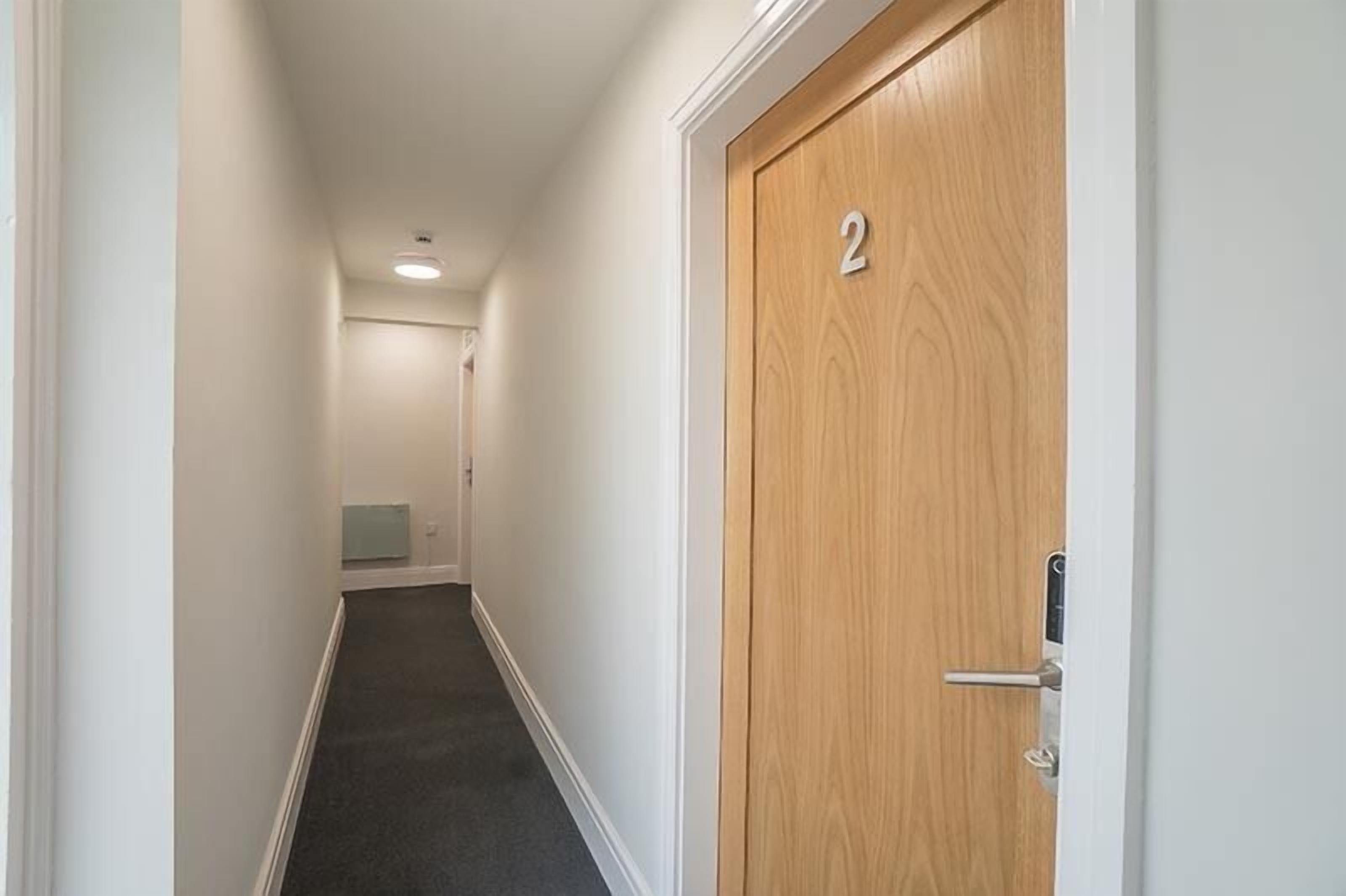 A narrow hallway with a wooden door marked with the number 2, a carpeted floor, and a light switch on the wall.