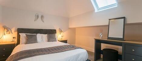A bedroom with a bed, bedside lamps, a desk, a mirror, and a skylight.