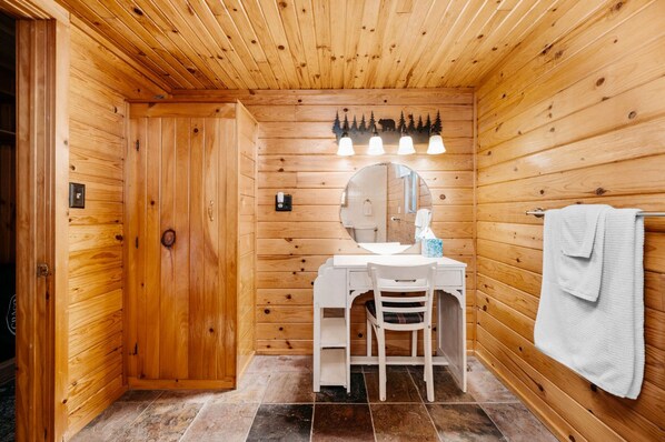A wooden cabin bathroom with a wooden door, a round mirror, a sink, a chair, and a towel rack.