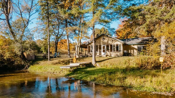 Une cabane rustique située au bord d’une rivière paisible, entourée d’arbres et d’une clôture en bois.