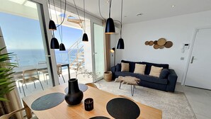 Ein modernes Wohnzimmer mit einem Holzesstisch, schwarzen Pendelleuchten, einem dunkelblauen Sofa und Blick auf den Ozean.