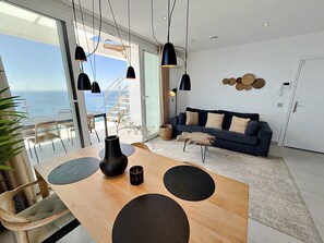 Una sala moderna con una mesa de comedor de madera, lámparas colgantes negras, un sofá azul oscuro y vista al océano.