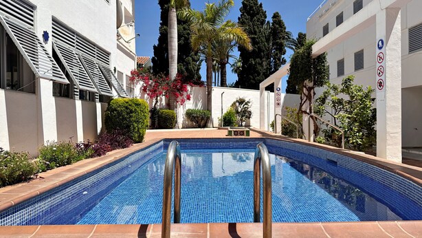 Una piscina con borde de baldosas, rodeada de edificios blancos con balcones y un cielo despejado.