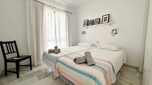 Un dormitorio con cama, una silla, una ventana con cortinas, una estantería con fotografías y una lámpara fijada a la pared.