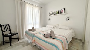 A bedroom with a bed, a chair, a window with curtains, a shelf with pictures, and a wall-mounted light.