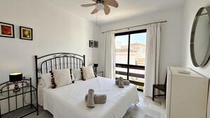 Une chambre à coucher avec un lit, un ventilateur de plafond, une fenêtre avec des rideaux, une table de chevet, un miroir et des tableaux encadrés accrochés au mur.