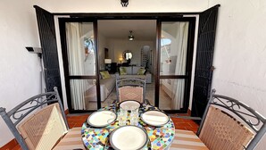Un patio abierto con una mesa de comedor preparada para dos, una silla de mimbre y vista hacia una sala de estar con sofá y una escalera.