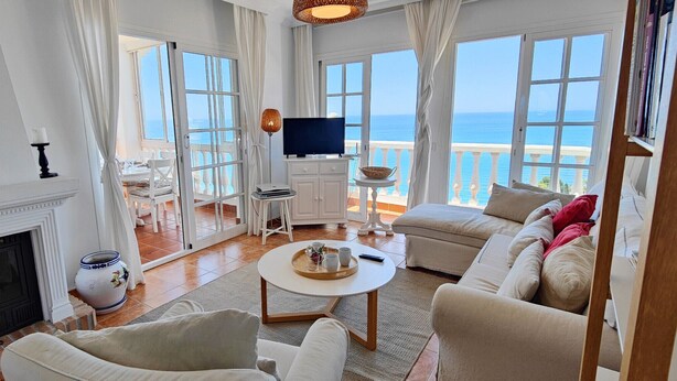 Amplia sala de estar con chimenea, sofá, mesa de centro y televisor. Amplias ventanas con vista al mar.