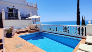 Una piscina con vista al mar, un edificio blanco y un balcón con barandilla.