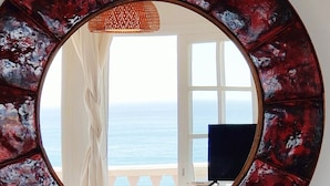 Un espejo redondo con un diseño rojo y negro, un tocador blanco con una vela y dos tazas estampadas, y vista al mar a través de una ventana.
