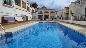 Une piscine dont les bords sont recouverts de carreaux, entourée d’une terrasse en bois et bordée de bâtiments blancs dotés de balcons.