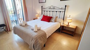 Un lit bien fait, recouvert d’une courtepointe à rayures, avec deux oreillers et un coussin rouge. On y trouve deux tables de chevet avec des lampes, une chaise et une fenêtre avec des rideaux.