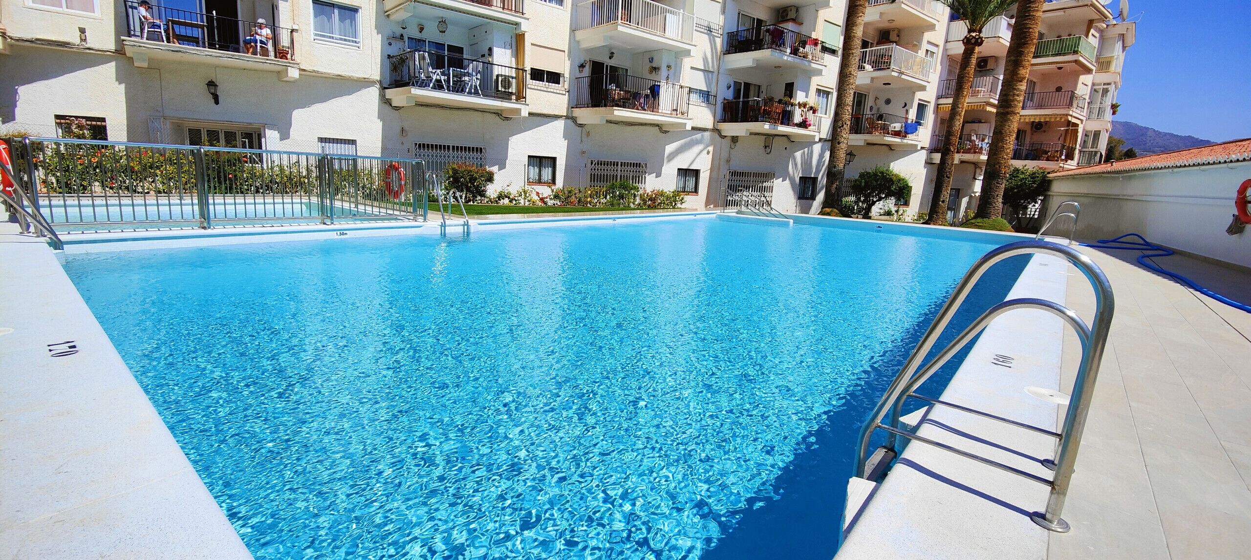 A clear blue swimming pool with a metal ladder, surrounded by a white building with balconies and palm trees.