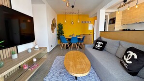 Un salón moderno con un televisor de pantalla plana, una mesa de centro de madera, un sofá gris con un cojín de Chanel y un comedor con sillas azules.