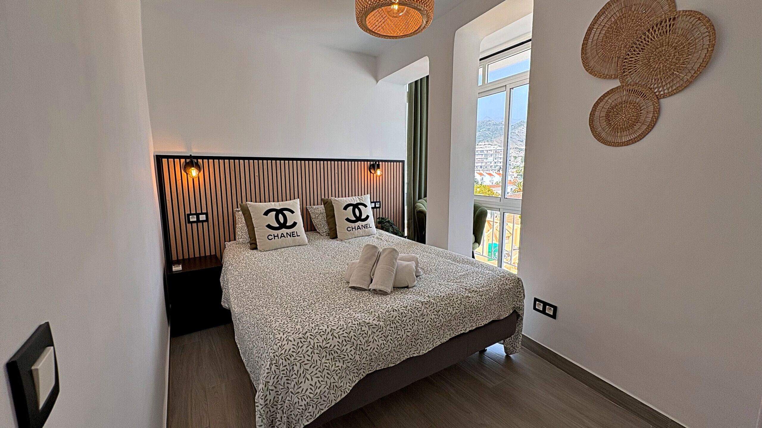 A neatly arranged bedroom with a bed, pillows, a bedside table, a hanging light fixture, and a window with a view.