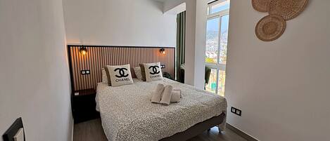 A neatly arranged bedroom with a bed, pillows, a bedside table, a hanging light fixture, and a window with a view.