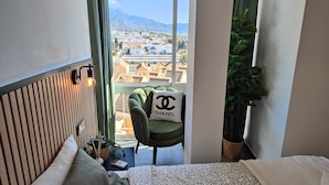 Habitación de hotel con cama, una silla con un cojín de Chanel, un ventanal con vistas a edificios y montañas, y una planta en maceta.