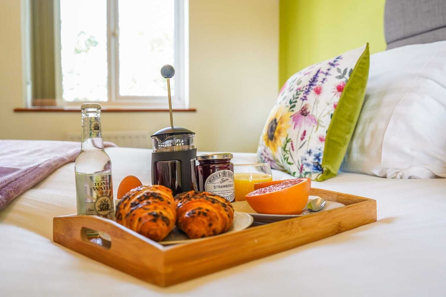 Ett brickbord med frukost placerat på sängen, med en flaska kolsyrat vatten, en kaffebryggare, ett glas jordgubbssylt, ett glas apelsinjuice, två croissanter och en halverad apelsin.