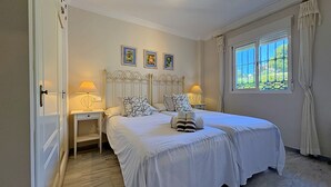 A bedroom with two beds, bedside tables with lamps, a window with curtains, and three framed pictures on the wall. - AP191 SEÑORIO DE CAPISTRANO (Nerja)