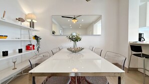 A modern dining area with a white table, clear chairs, a central vase with flowers, a ceiling fan, and a kitchen area with a coffee maker.
