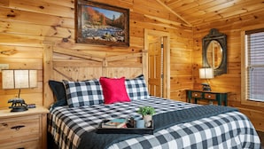 Et soveværelse i en træhytte med et ternet sengetæppe, en rød pude, et natbord med en lampe, et indrammet billede på væggen og et vindue med persienner.