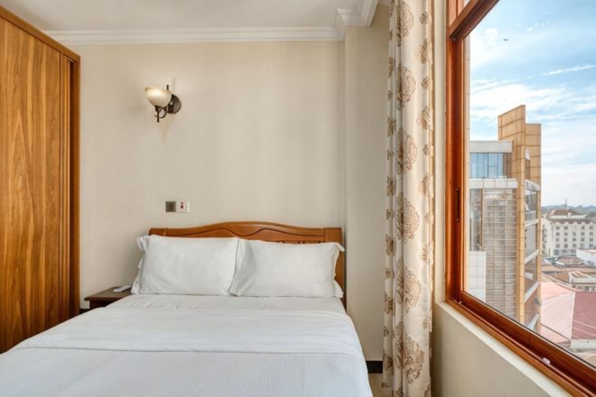 A hotel room with a bed, wooden headboard, a nightstand, a wall-mounted light fixture, and a window with a city view.